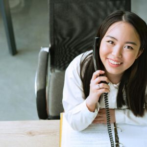 Smiling woman in a white shirt talking on the phone while sitting at an office desk.