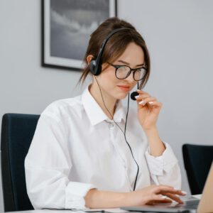 Young woman in glasses and headset providing customer support at a laptop in an office setting.